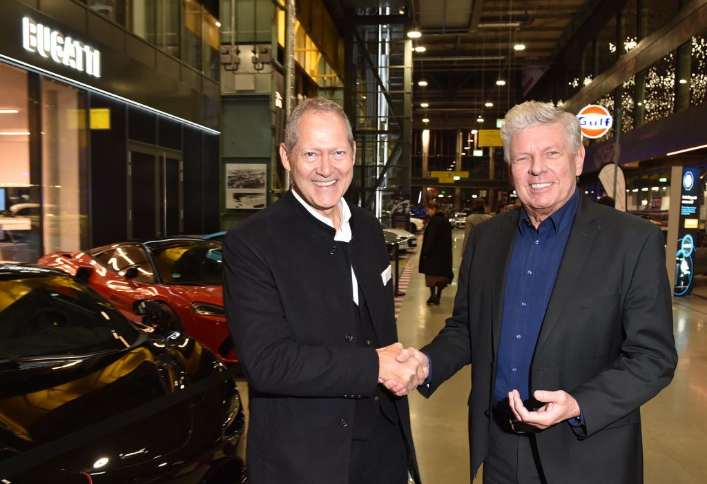 Zwei Männer begrüßen sich mit Handschlag in einer Eventlocation mit Luxusautos und Bugatti-Logo im Hintergrund