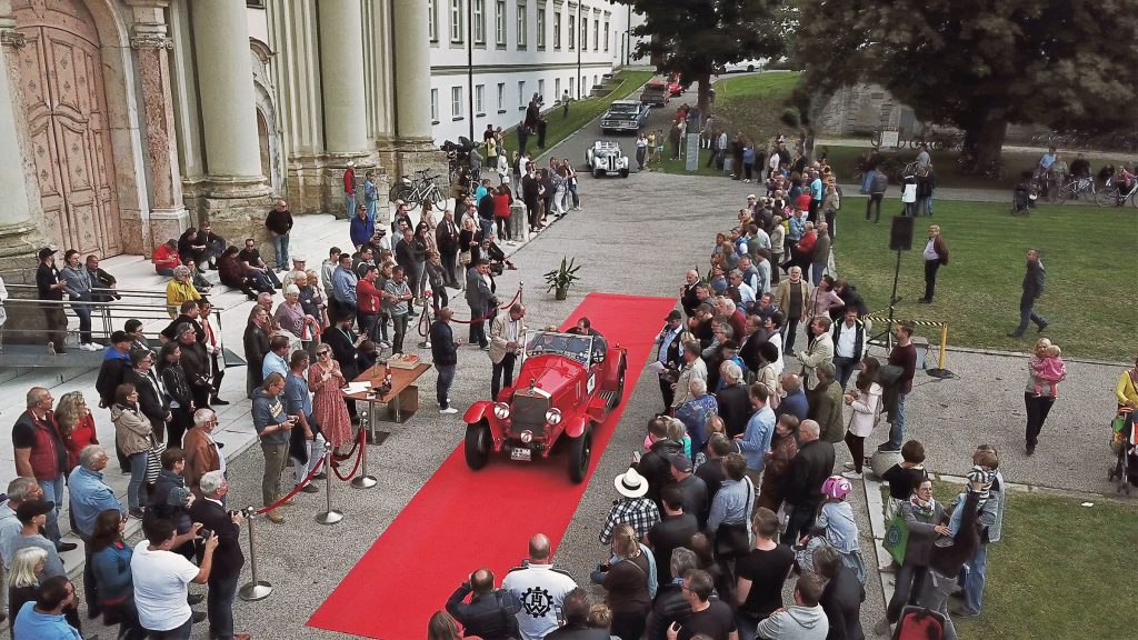 Klassische Oldtimer fahren über roten Teppich bei einem Event vor historischem Gebäude mit großem Publikum