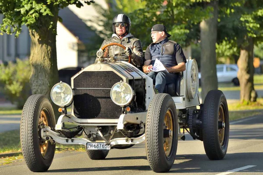 Zwei Männer fahren in einem historischen Oldtimer bei einem Event auf einer sonnigen Straße mit Bäumen im Hintergrund