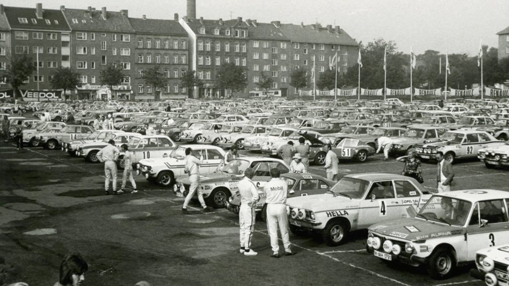 Historisches Motorsport-Event mit zahlreichen Rennfahrzeugen und Fahrern auf einem großen Parkgelände vor Wohnhäusern