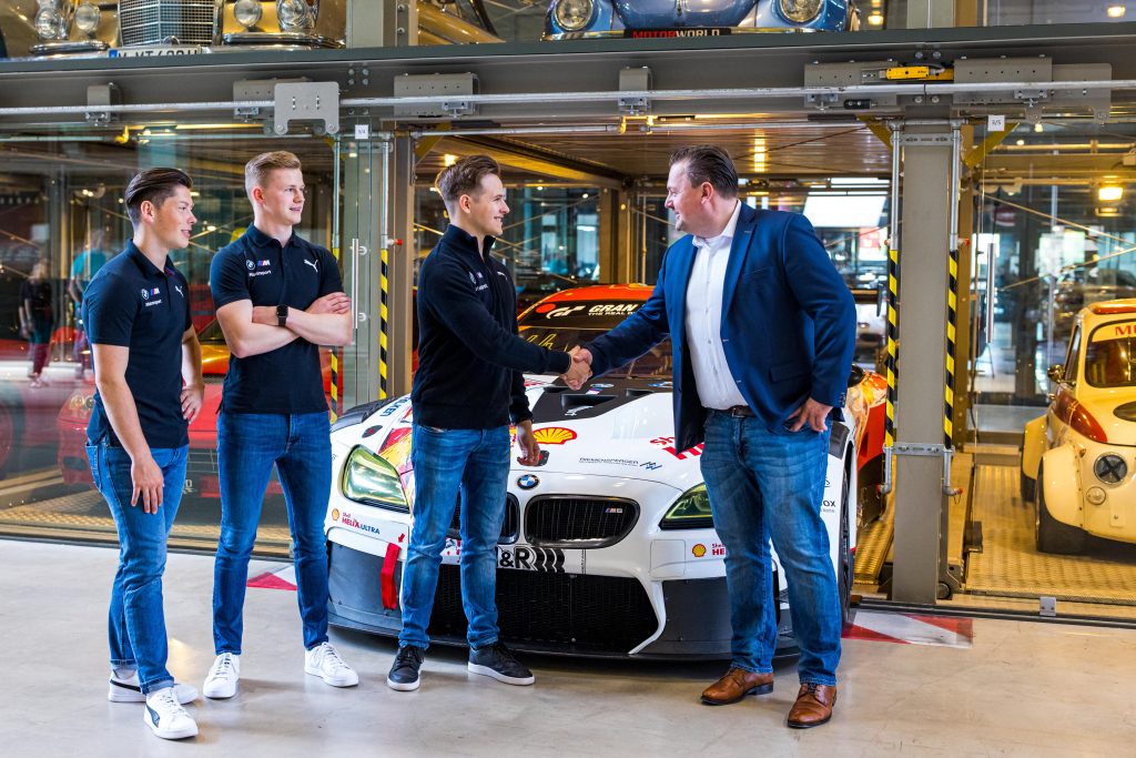 Junge Männer in Sportbekleidung begrüßen Geschäftsmann in Eventlocation mit Motorsport-Autos im Hintergrund