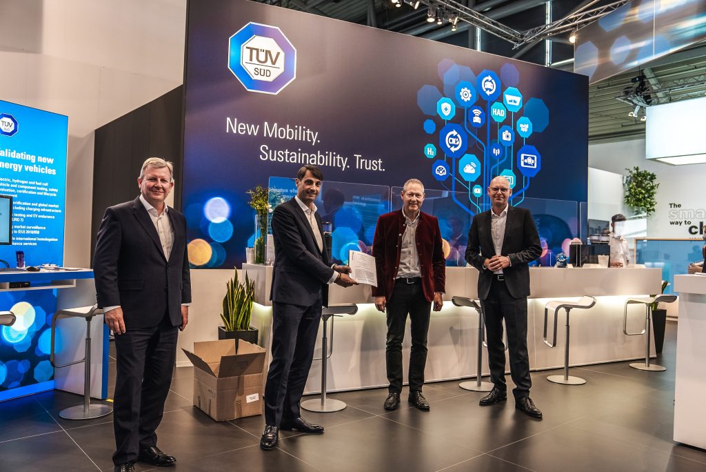Vier Männer in Anzügen auf einem Event vor einem TÜV Süd Messestand mit blauer digitaler Grafik