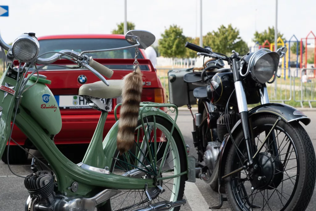 Zwei klassische Motorräder und ein rotes BMW-Auto auf einem Parkplatz bei einem Event
