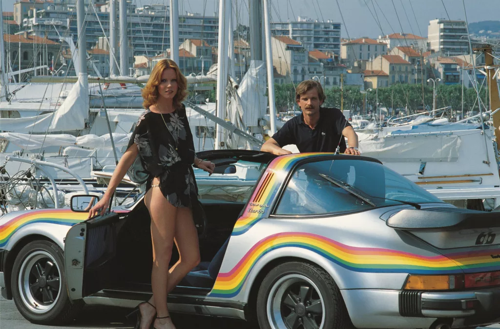 Frau und Mann mit silbernem Porsche 911 Targa Turbo mit Regenbogenstreifen vor Marina mit Segelbooten