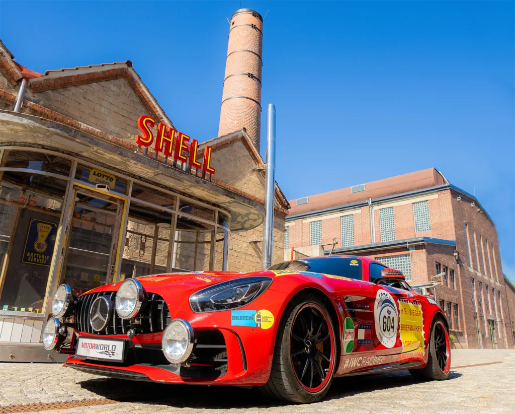 Roter Rennsportwagen vor historischer Shell-Tankstelle in einer Industrie-Eventlocation bei sonnigem Wetter