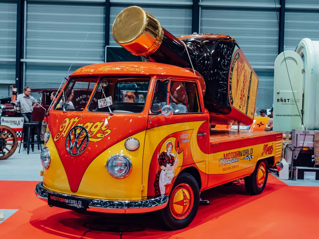 Retro VW-Bus in Gelb und Rot mit einer riesigen Maggi-Flasche auf dem Dach bei einem Event in einer Halle
