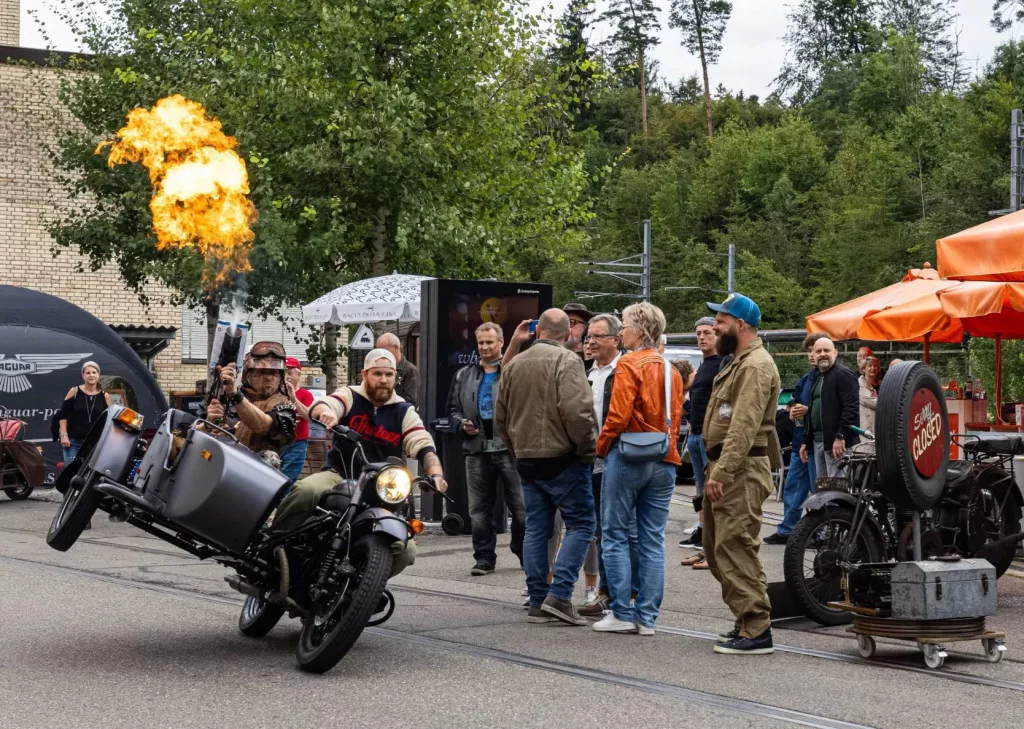 Event mit Motorrad und Seitenwagen, Feueraktion und Zuschauern vor grüner Baumkulisse und Gebäuden