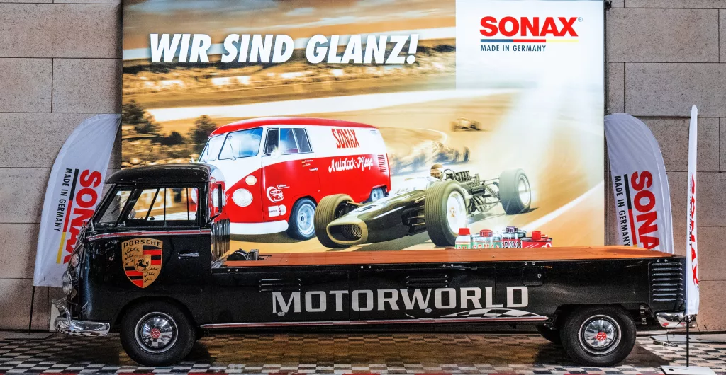 Schwarzer Motorworld Transporter vor einer Werbewand mit Sonax Event-Motiven und alten Rennfahrzeugen