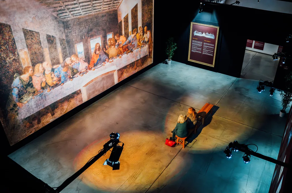 Zwei Personen sitzen auf einer Bank in der Eventlocation vor einem beleuchteten Gemälde des Letzten Abendmahls in der Ausstellung