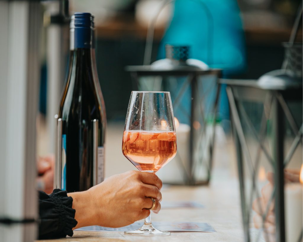 Person hält ein Glas Roséwein neben einer Weinflasche bei einem entspannten Event