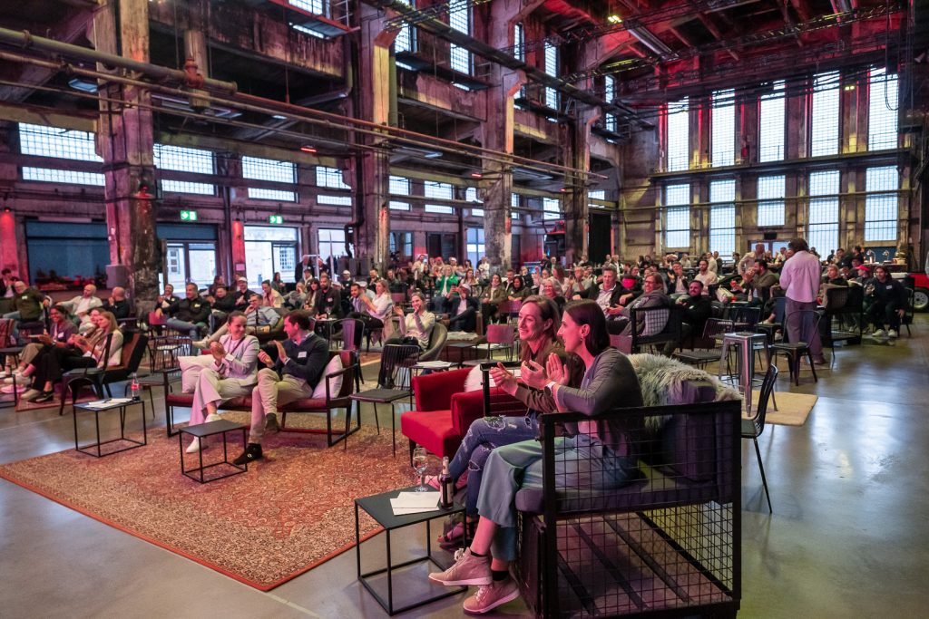 Große Eventlocation mit Publikum bei einer Veranstaltung in einer industriellen Halle mit Lounge-Möbeln und bunter Beleuchtung