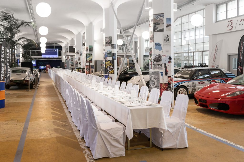 Elegante Eventlocation mit langem, weiß gedecktem Tisch neben klassischen und Sports cars in moderner Halle