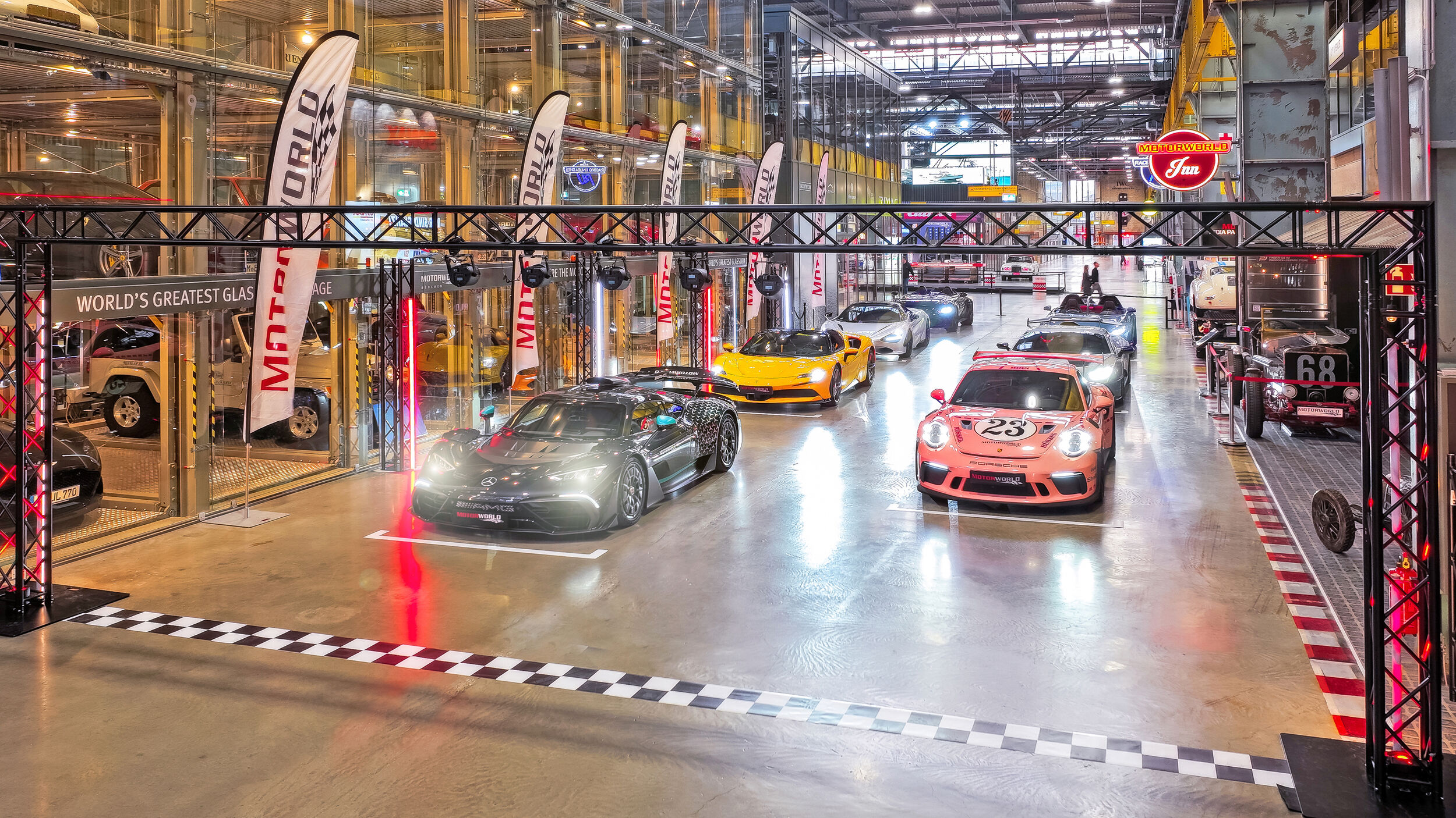 Moderne Supersportwagen in einer hell beleuchteten, großen Ausstellungshalle mit Glasfassade und Motorworld-Bannern, unter einem Rennstartbogen mit schwarz-weiß kariertem Bodenmuster.