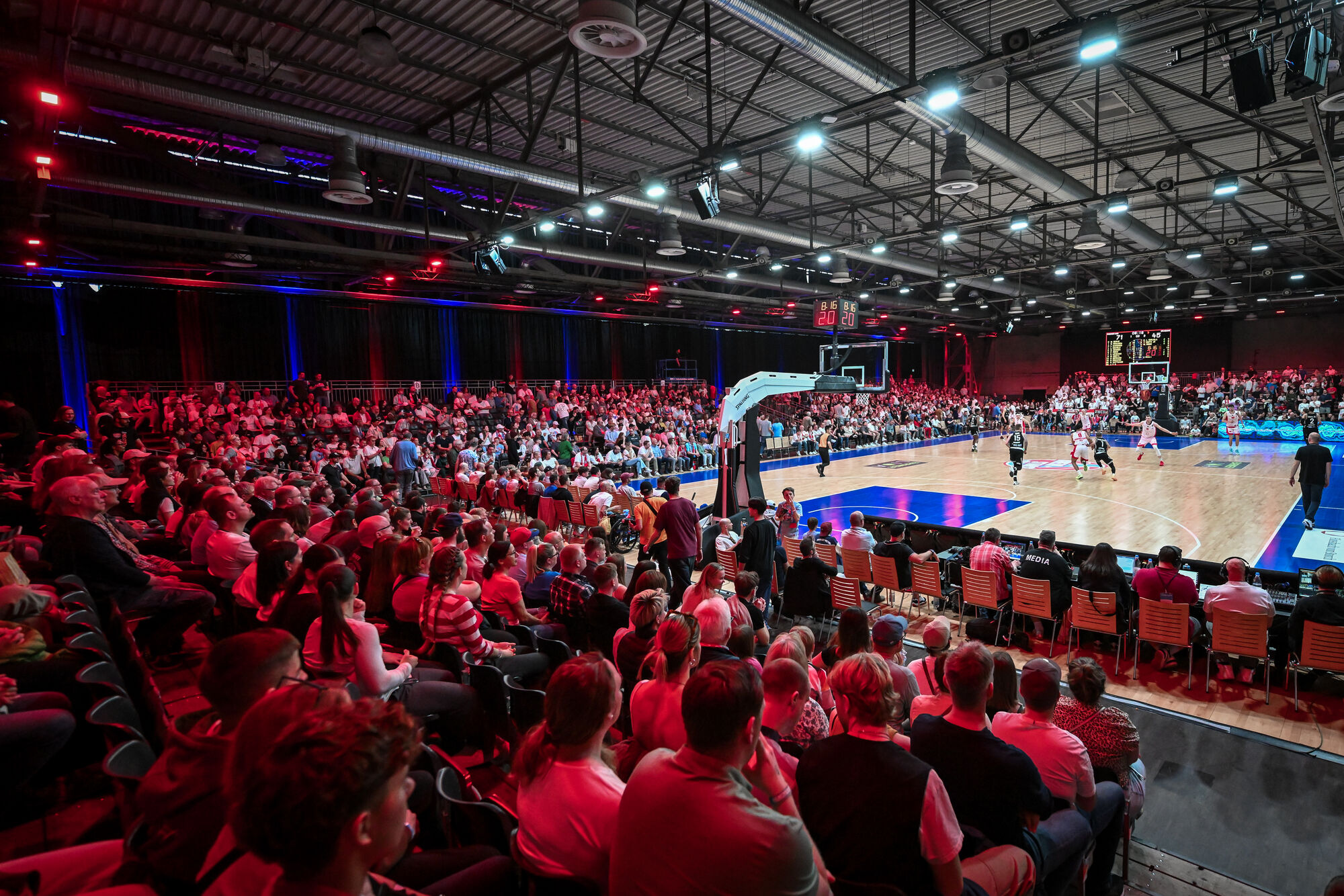 Blick auf eine gut besuchte Basketballhalle mit Zuschauern auf Tribünen und Tribünenrängen, die einem Basketballspiel folgen, das auf einem gut beleuchteten Spielfeld stattfindet.