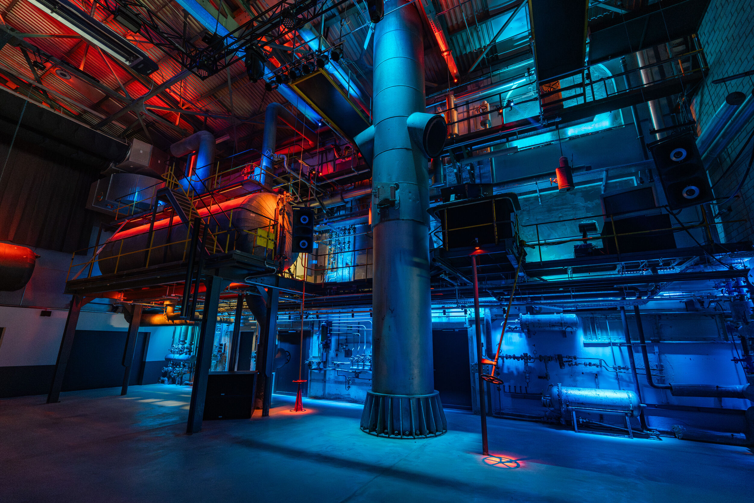 Industrielle Anlage mit großen Rohren und Metallkonstruktionen, beleuchtet in Blau und Rot, mit sichtbaren Ventilen und Leitungen in einer Halle mit Betonboden und hoher Decke.