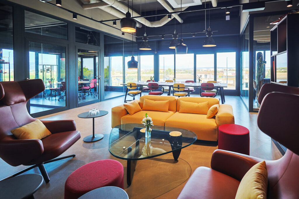 Moderner Besprechungsraum mit gelbem Sofa, roten Sesseln, bunten Stühlen und großem Tisch vor Panorama-Fenstern, die eine Landschaft zeigen.