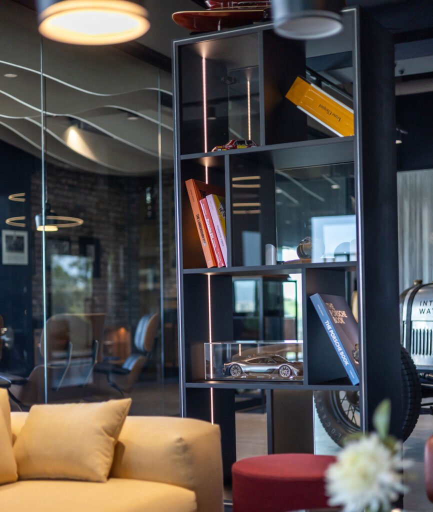 Moderner Wohnraum mit einem schwarzen Regal, das Bücher, ein Modellauto und eine gelbe Mappe enthält, im Vordergrund ein gelbes Sofa und ein roter Hocker, im Hintergrund eine Glaswand mit reflektierenden Lichtern und einem alten Auto erkennbar.