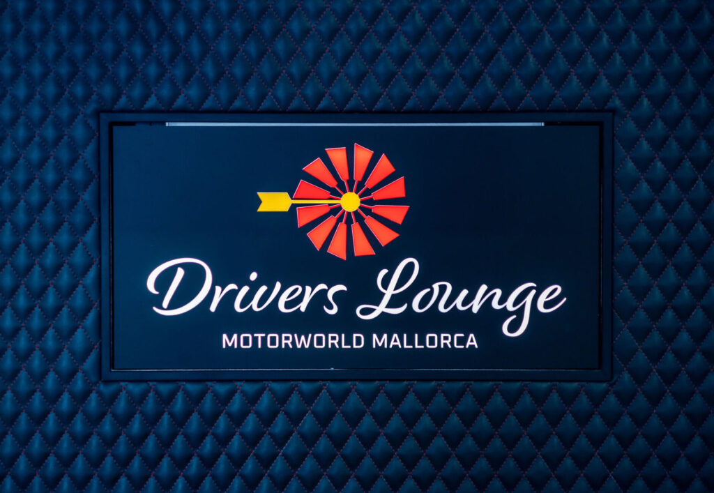 Leuchtschild mit dem Schriftzug "Drivers Lounge MOTORWORLD MALLORCA" vor einer dunkelblauen, gesteppte Polsterung.