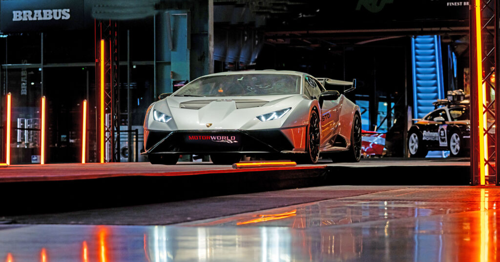 Weißer Lamborghini Huracán STO vor dunklem Hintergrund mit roten Neonlichtern und reflektierendem Boden, davor steht ein schwarzes Polizeiauto mit Aufschrift.