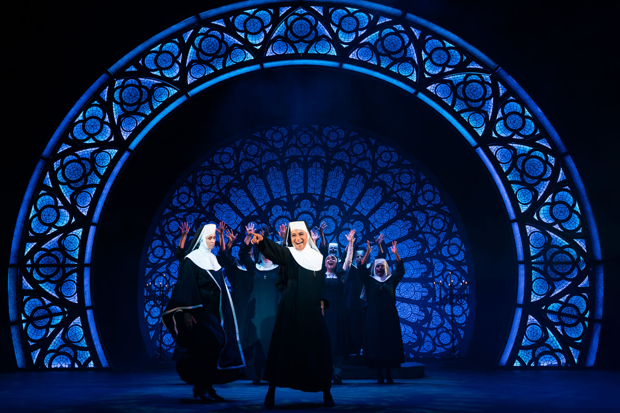 Theaterbühne mit mehreren als Nonnen verkleideten Darstellern vor einem großen blauen Fenster mit gotischem Rosettenmuster, die in einer lebhaften Pose mit erhobenen Armen stehen, wobei die vorderste Nonne nach vorne zeigt und lächelt.