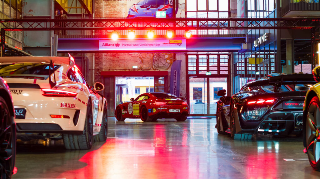 Innenaufnahme einer Garage oder eines Autohauses mit sportlichen Luxusautos, vor allem Porsche und Mercedes AMG, bei dem roten und warmen Lichtreflexen auf glänzendem Boden.