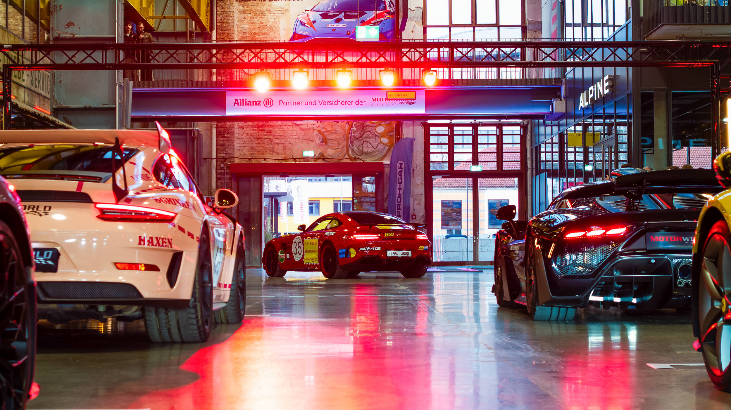 Innenaufnahme einer Garage oder eines Autohauses mit sportlichen Luxusautos, vor allem Porsche und Mercedes AMG, bei dem roten und warmen Lichtreflexen auf glänzendem Boden.
