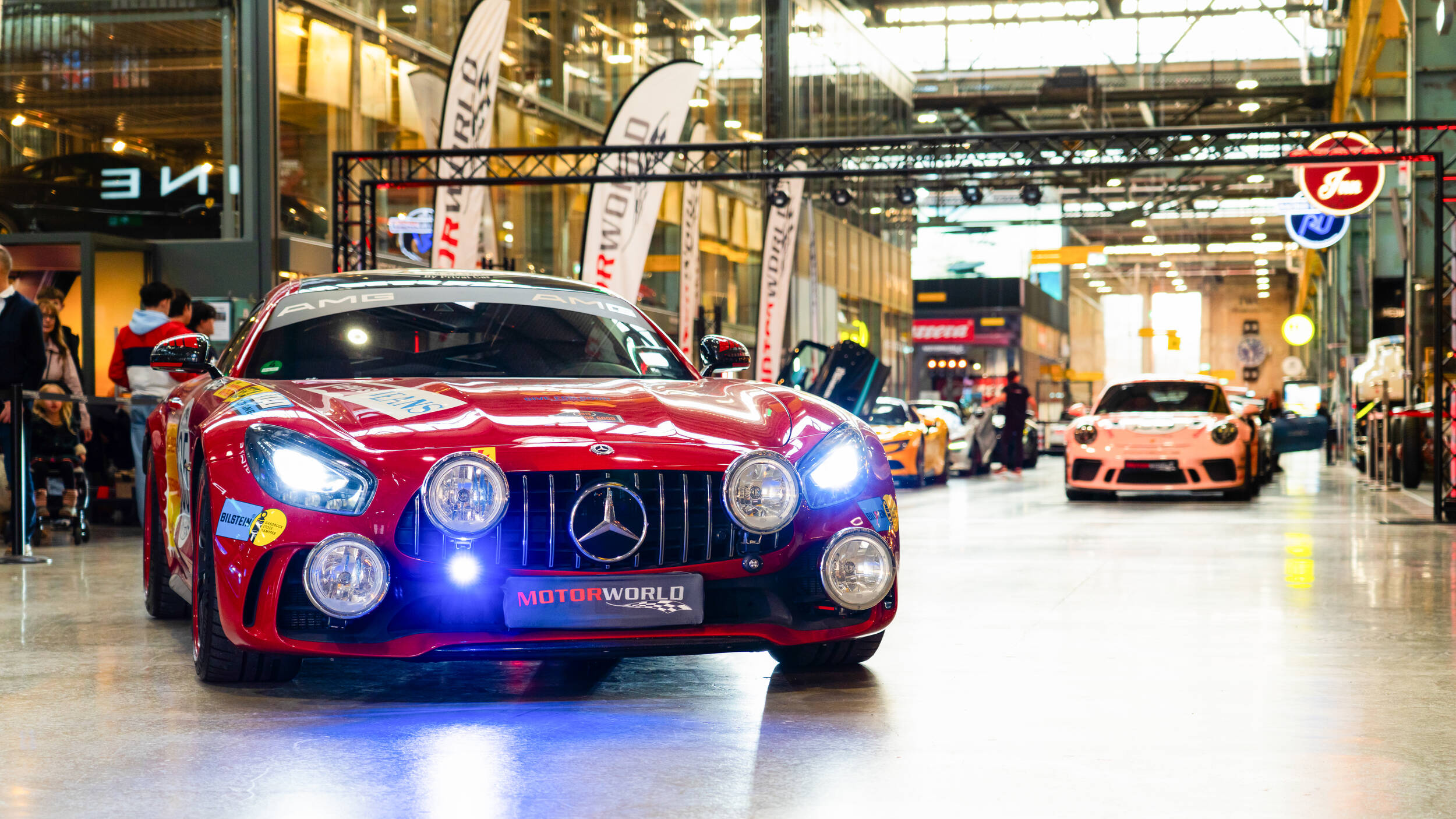 Roter Mercedes-AMG Rennwagen mit hellen Scheinwerfern und zusätzlichen Frontscheinwerfern in einer hell erleuchteten Halle mit weiteren Sportwagen und Personen im Hintergrund.