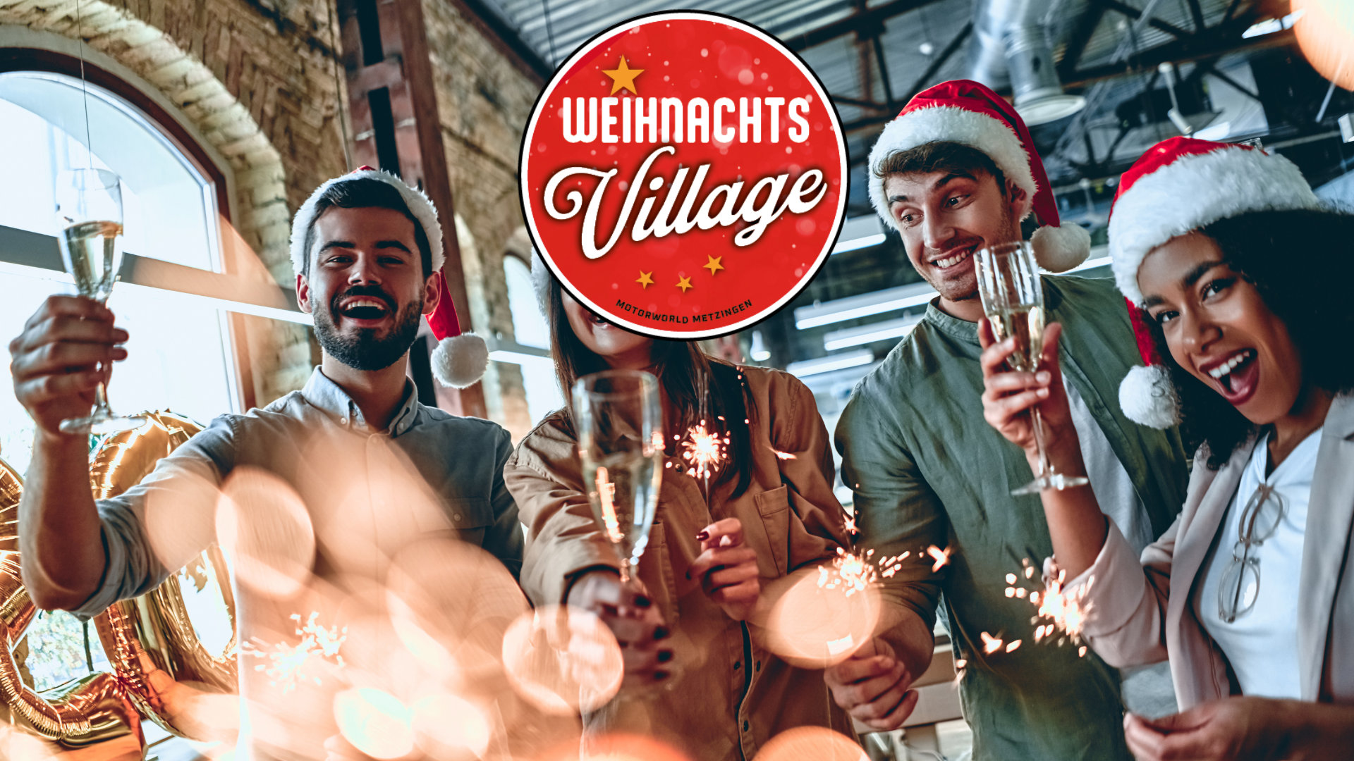 Vier junge Erwachsene in Weihnachtsmützen feiern mit Sektgläsern und Wunderkerzen in einem modernen Raum mit großen Fenstern und Industriecharakter, darüber ein rotes rundes Logo mit der Aufschrift Weihnachts Village Motorworld Metzingen.