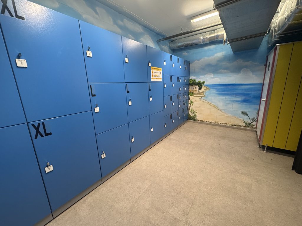 Ein Raum mit blauen und gelben Schließfächern, an der Wand ein Wandbild mit einer Strand- und Meeresszenerie unter einem wolkigen Himmel, beleuchtet durch Deckenleuchten.