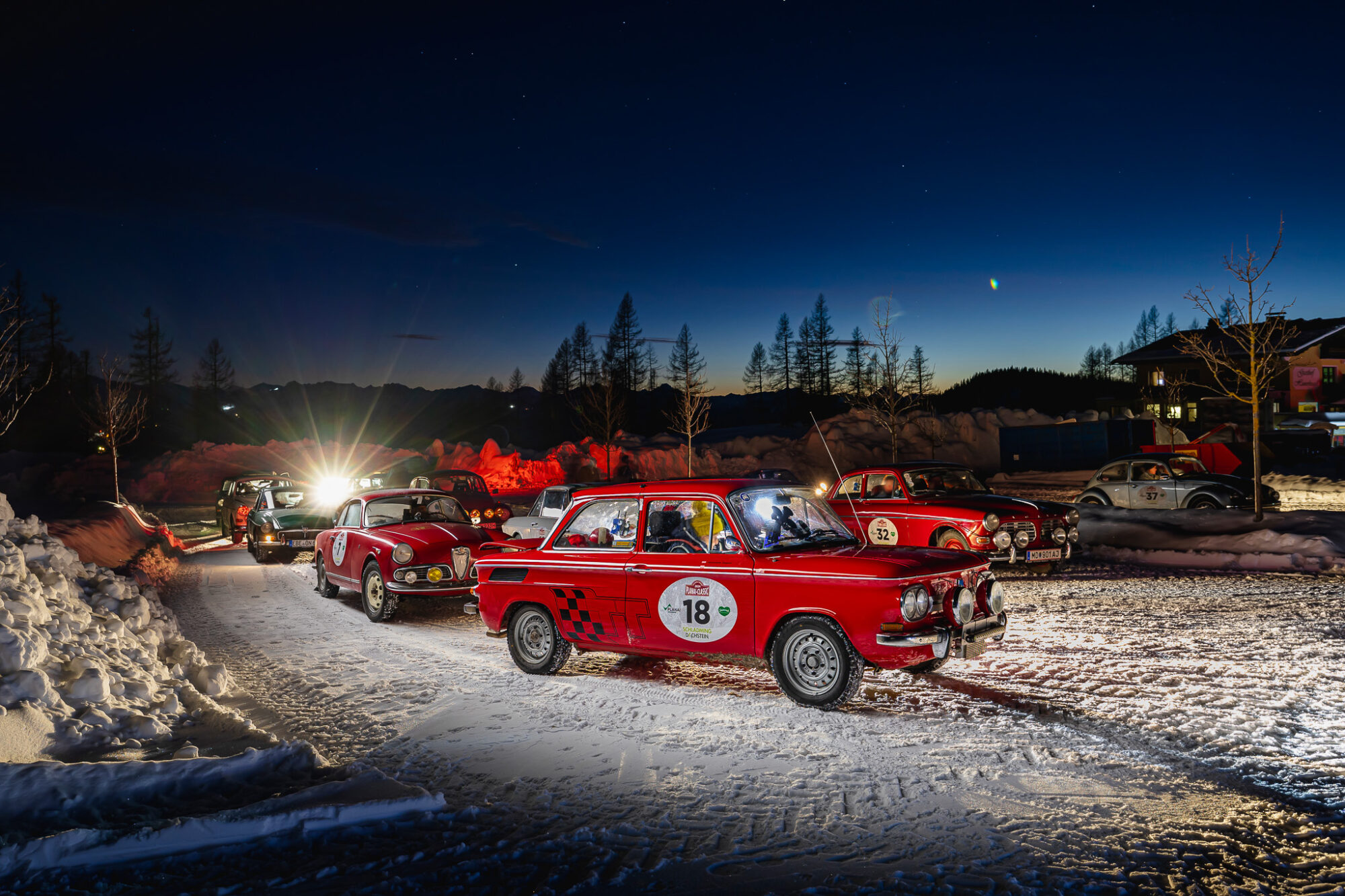 Reihe von klassischen roten und weißen Oldtimerrallye-Autos auf schneebedeckter Straße bei Nacht vor einem bewaldeten Bergpanorama mit klarem Sternenhimmel.