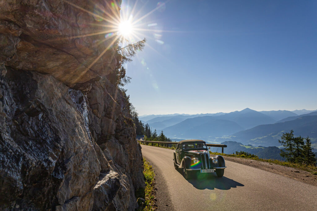 Alte Cabrio-Automobil auf Bergstraße bei sonnigem Himmel mit Bergpanorama im Hintergrund und Felsen an der linken Seite.