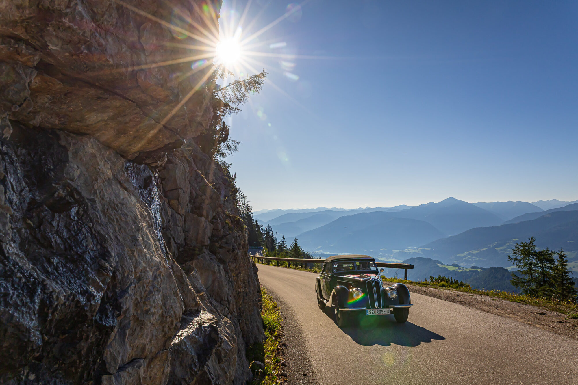 Alte Cabrio-Automobil auf Bergstraße bei sonnigem Himmel mit Bergpanorama im Hintergrund und Felsen an der linken Seite.