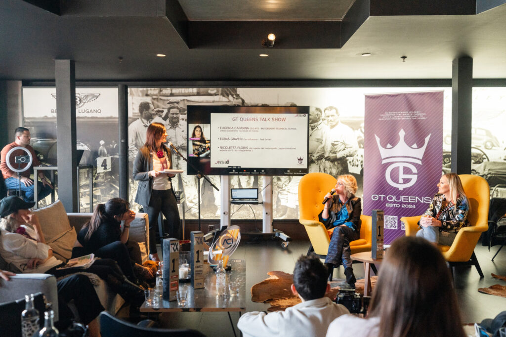 Frauen-Podiumsdiskussion bei der GT Queens Talk Show mit einer Moderatorin stehend neben einem Bildschirm, zwei Frauen in gelben Sesseln mit Mikrofonen, Publikum im Vordergrund und großem GT Queens-Banner im Hintergrund.