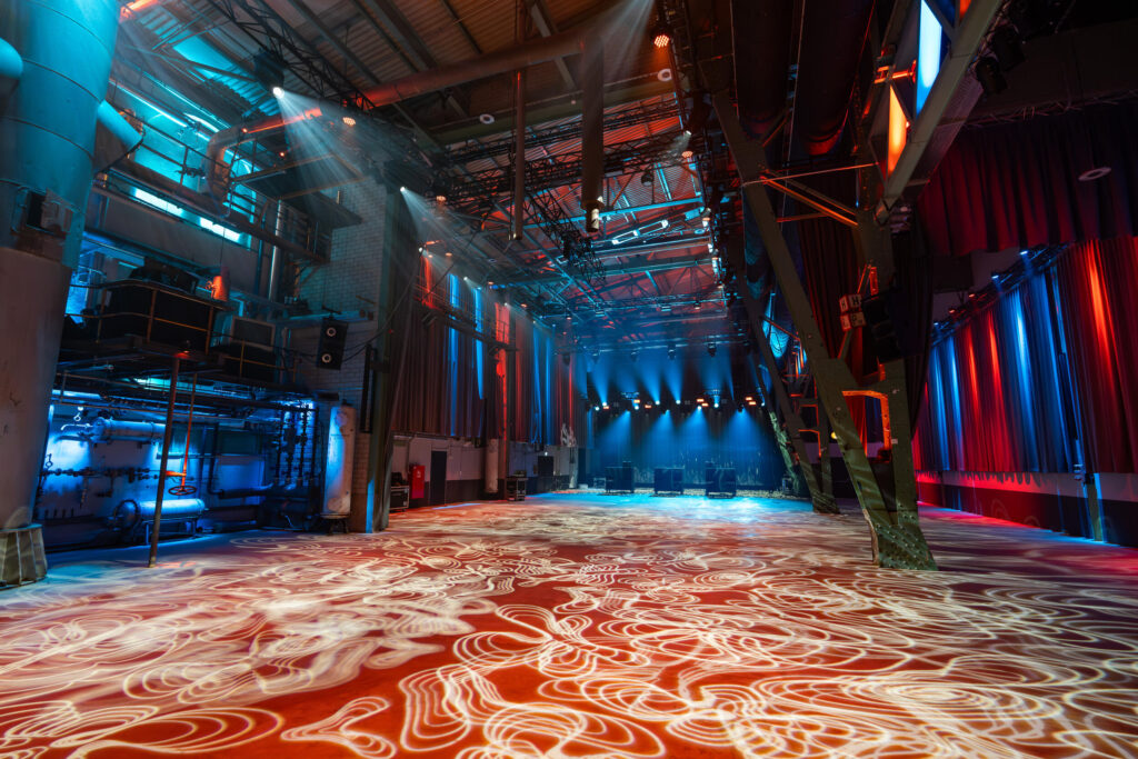 Großer, leerer Veranstaltungssaal mit hohen Decken, Industriekonstruktionen und bunter, atmosphärischer Beleuchtung in Blau und Rot, mit Lichtmustern auf dem Boden und Scheinwerfern an der Decke.