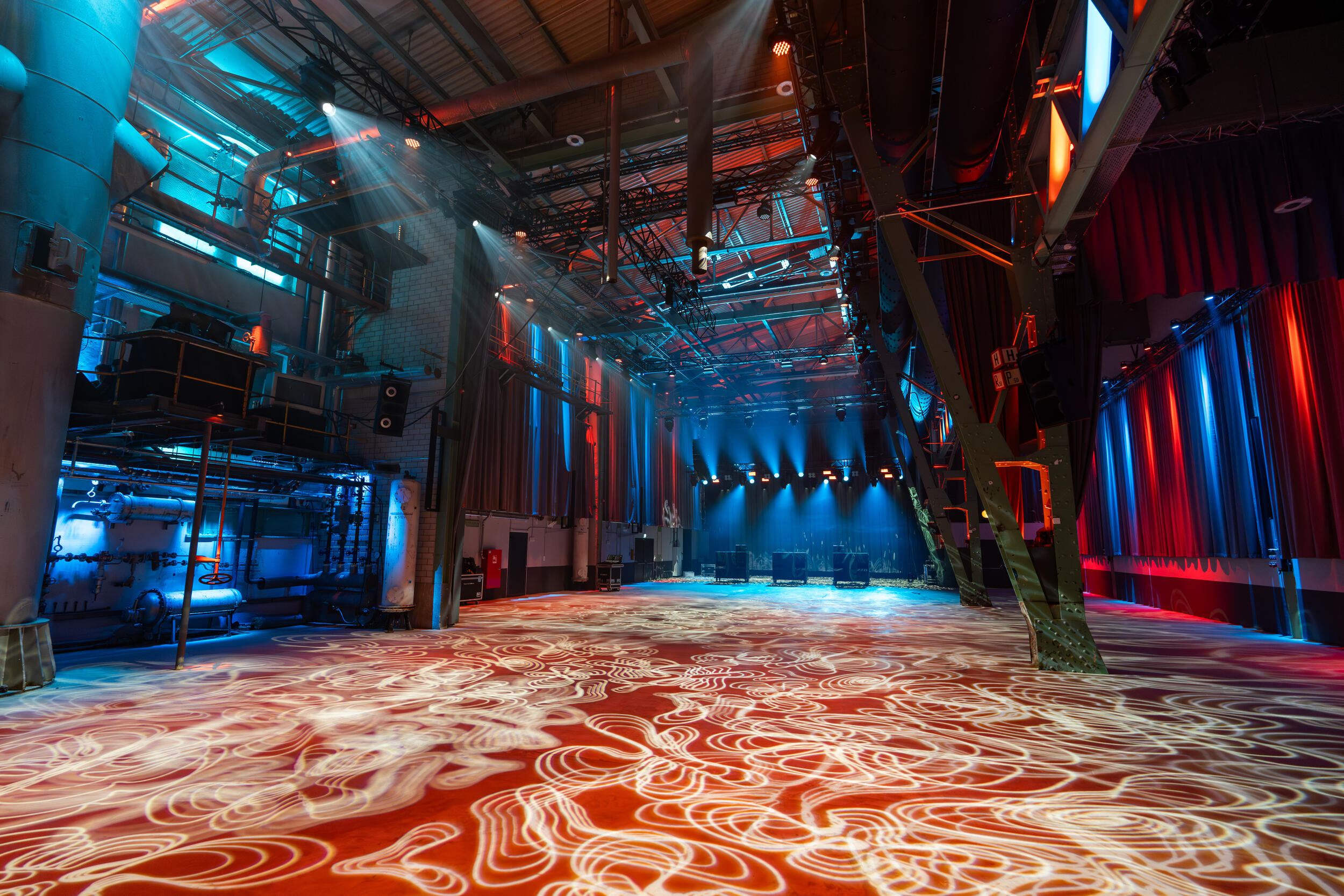 Großer, leerer Veranstaltungssaal mit hohen Decken, Industriekonstruktionen und bunter, atmosphärischer Beleuchtung in Blau und Rot, mit Lichtmustern auf dem Boden und Scheinwerfern an der Decke.