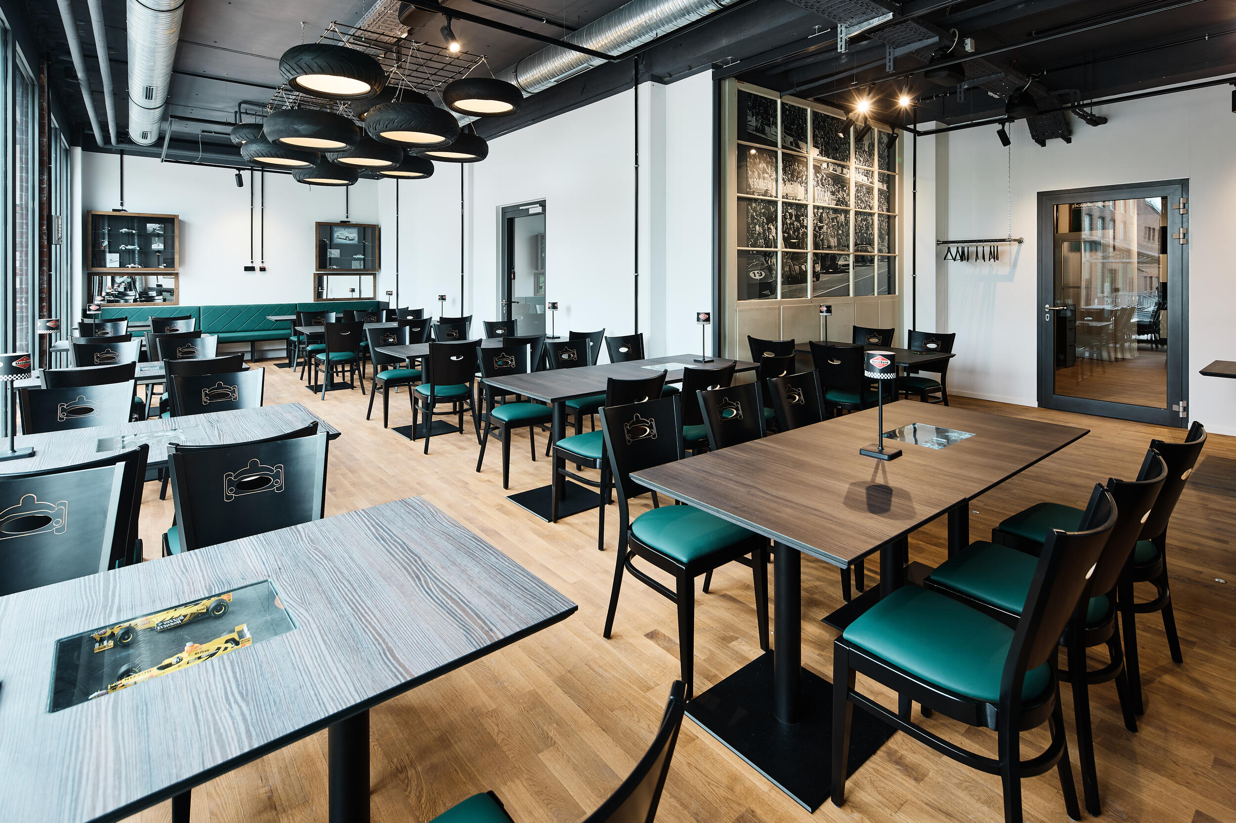 Moderner Gastraum eines Restaurants mit schwarzen Stühlen mit grünem Sitzpolster, Holztischen mit eingelassenen Modellautos, großen Deckenlampen aus Autoreifen und einer Wand mit Fotocollage historischer Rennen.
