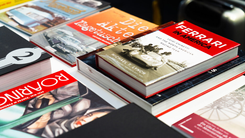 Nahaufnahme eines Tischs mit mehreren übereinander gestapelten Büchern, darunter Titel wie »Ferrari in America« und »Roaring« mit Bildern von Autos und historischen Fotos auf den Buchcovern.