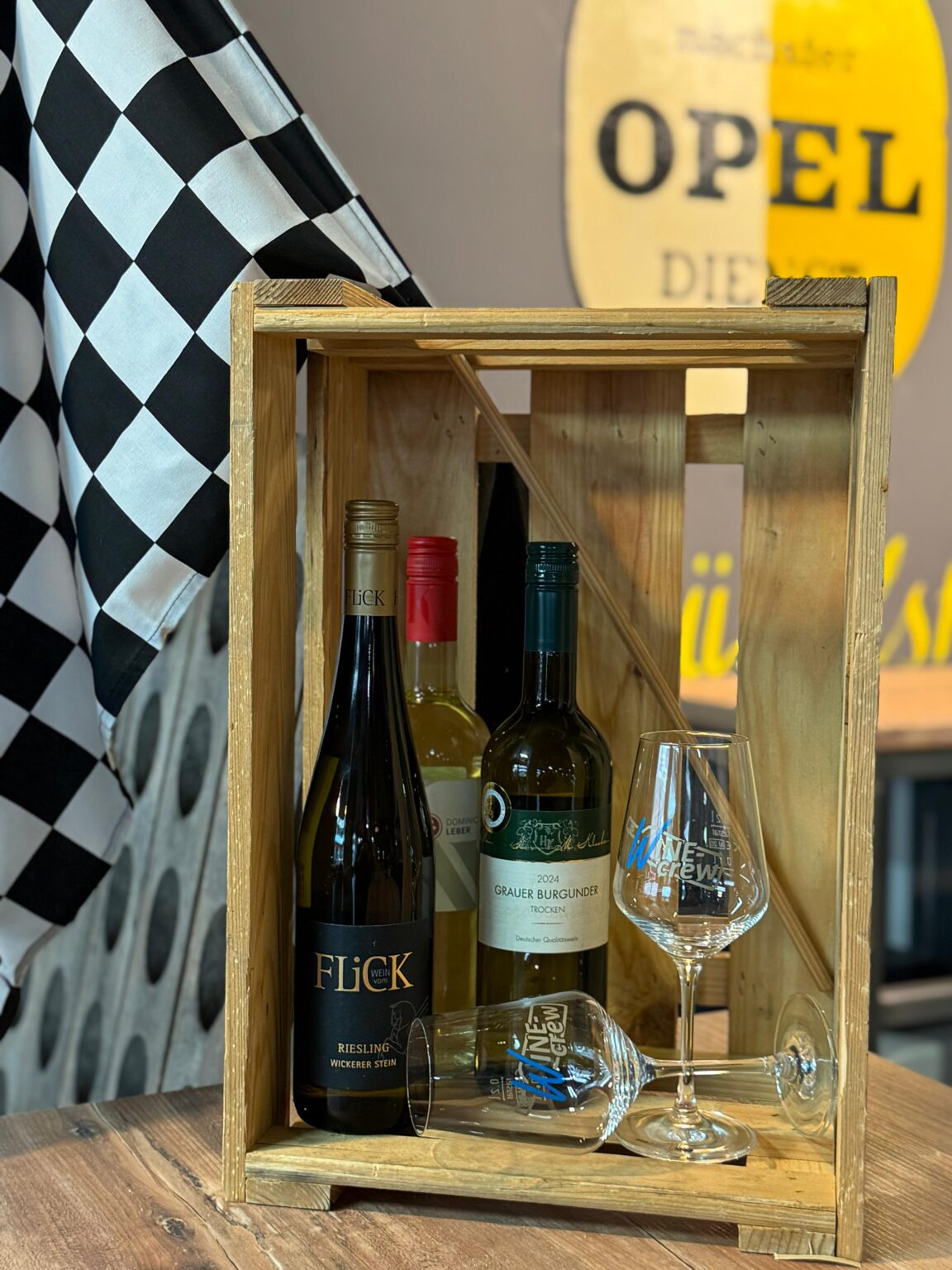 Drei Weinflaschen in einem Holzrahmen auf einem Holztisch, zwei Weingläser mit Aufschrift "WINE crew", eine schwarz-weiß karierte Flagge im Hintergrund und eine gelbe Opel-Werbung an der Wand.