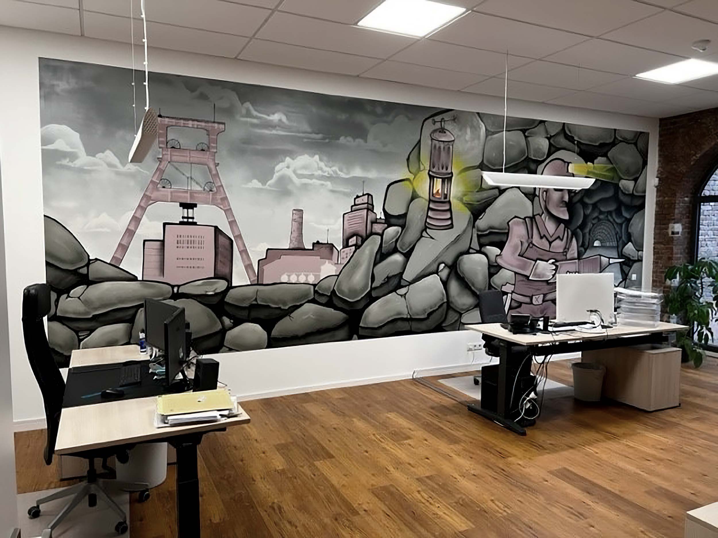 Büro mit zwei Schreibtischen und Stühlen vor einer großen Wand mit einem Wandgemälde, das eine Bergbau-Szene mit großen Steinen, einer Bergbaustätte und einem Bergmann mit Grubenlampe zeigt.