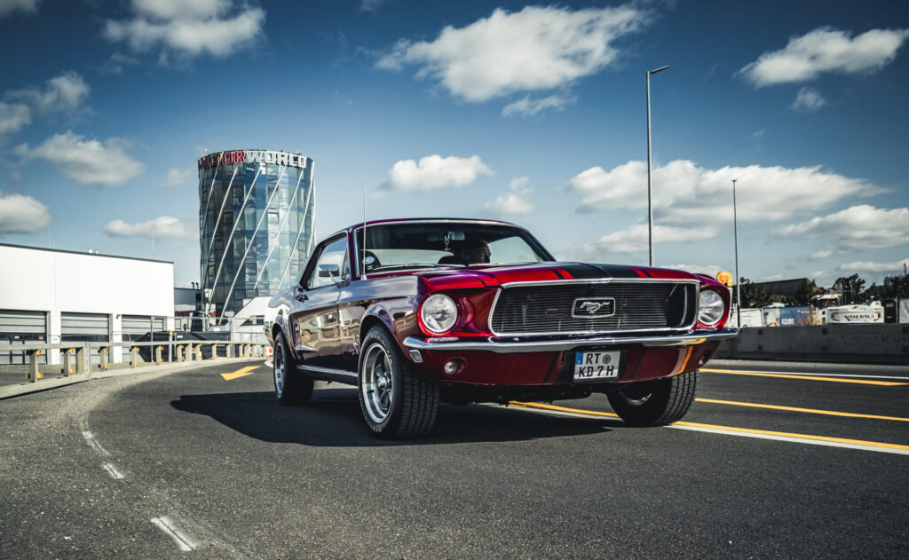 Roter klassischer Ford Mustang mit schwarzen Rennstreifen auf einer Straße vor einem modernen Gebäude mit der Aufschrift Motorworld bei blauem Himmel mit Wolken.