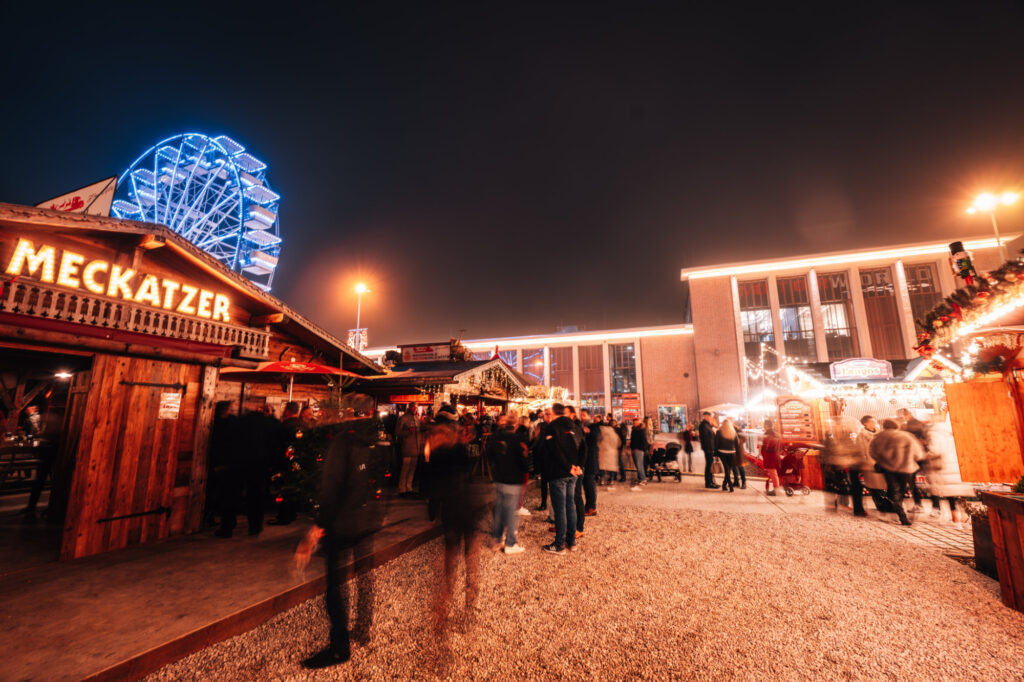 Abendaufnahme eines beleuchteten Weihnachtsmarktes mit Menschen, Holzständen, einem beleuchteten Riesenrad im Hintergrund und Gebäude mit festlicher Beleuchtung.