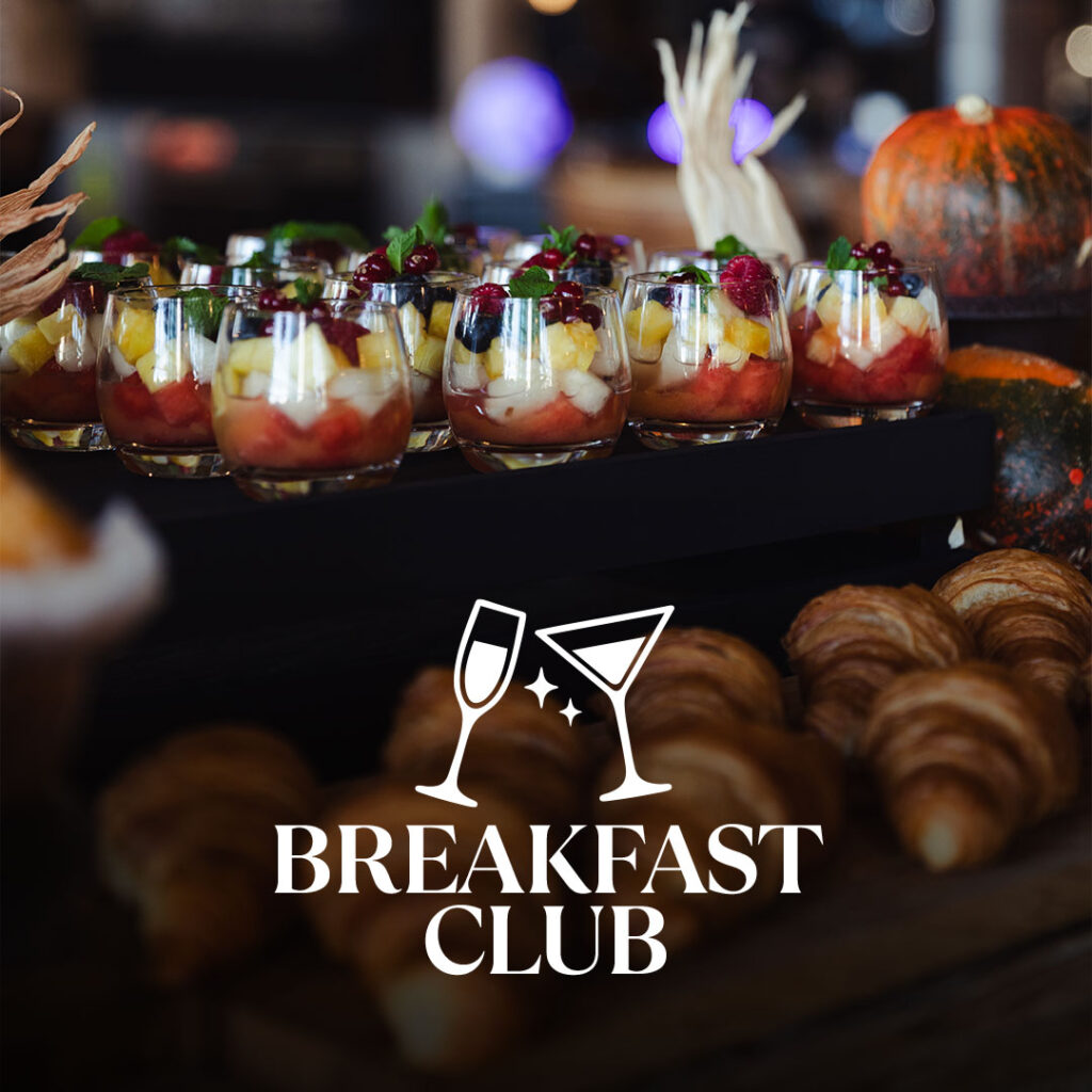 Verschiedene kleine Gläser mit Obstsalat und frischen Beeren auf einem Tablett, davor liegen mehrere Croissants, im Hintergrund liegen Kürbisse, darüber der Schriftzug 'Breakfast Club' mit Symbolen von zwei anstoßenden Gläsern.