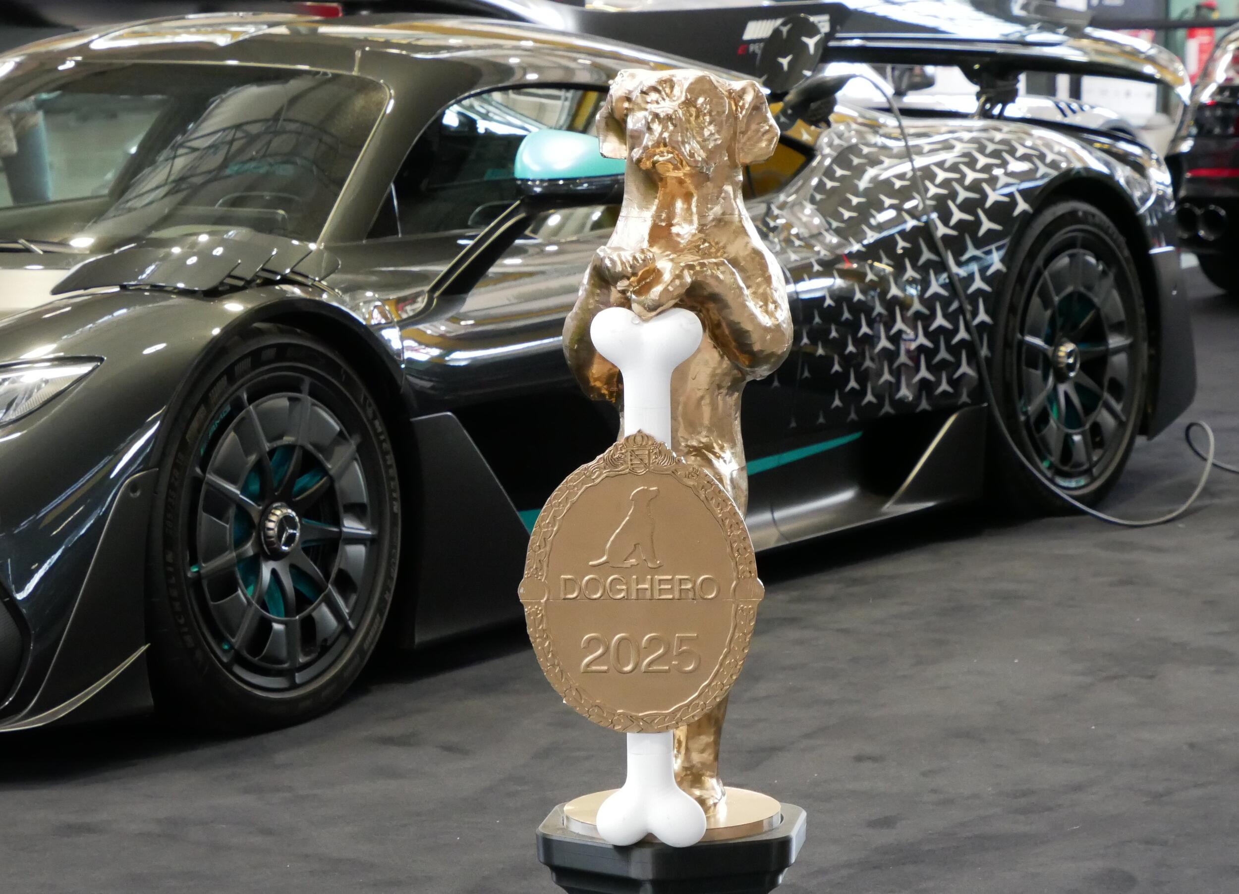 Goldene Hundestatue hält einen Knochen mit einer Plakette, auf der DOGHERO 2025 steht, vor einem schwarzen Sportwagen mit Muster und Ladekabel in einer Ausstellungshalle.