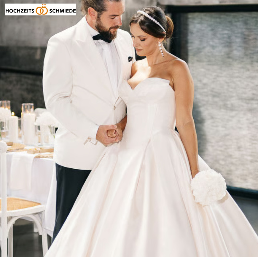Bräutigam im weißen Smoking mit schwarzer Fliege und Braut in weißem trägerlosem Brautkleid mit Perlenhaarreif und großen Perlenohrringen, die sich an den Händen halten, im Hochzeitsambiente mit Kerzen und Tischdekoration im Hintergrund