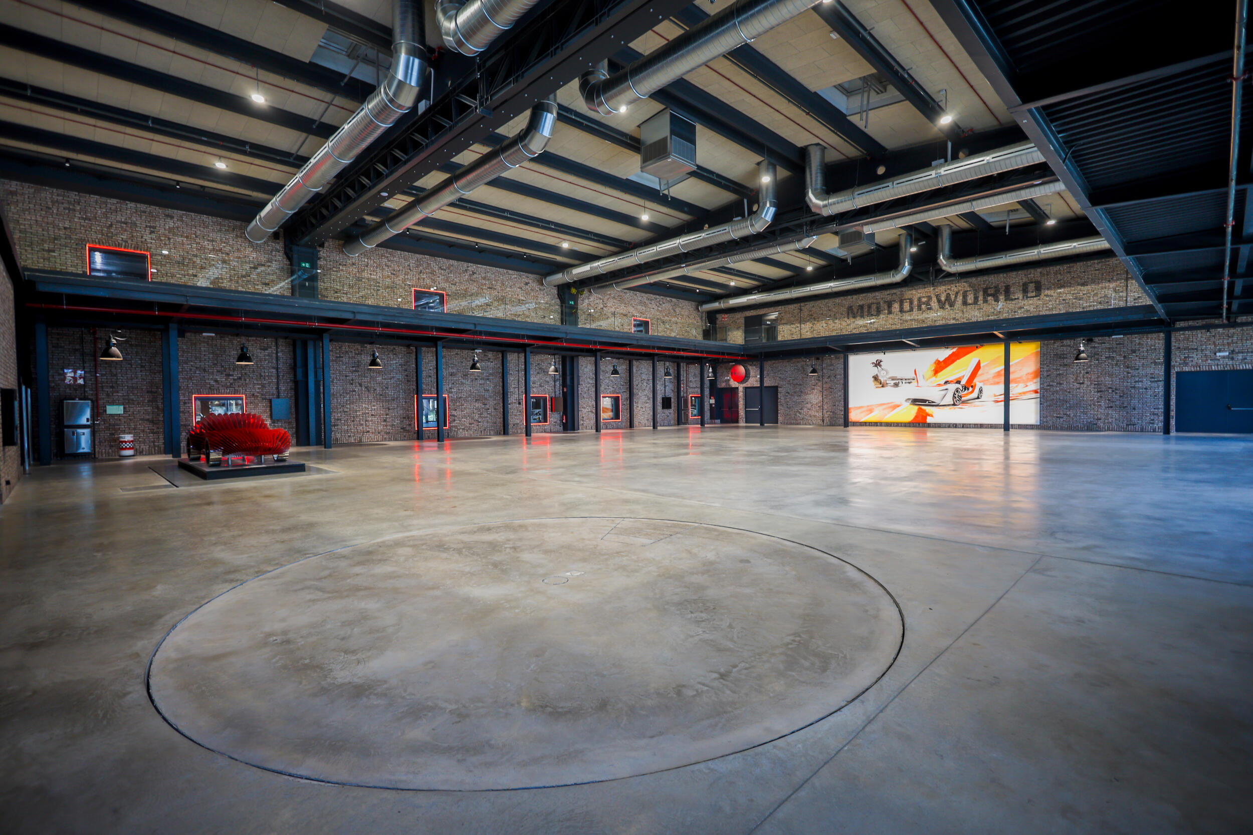 Große Industriehalle mit sichtbaren Backsteinwänden, Rohren an der Decke und poliertem Betonboden mit großem Kreis markiert, roter Skulptur und großem Wandbild eines Autos im Hintergrund mit Schriftzug "MOTORWORLD" an der Wand
