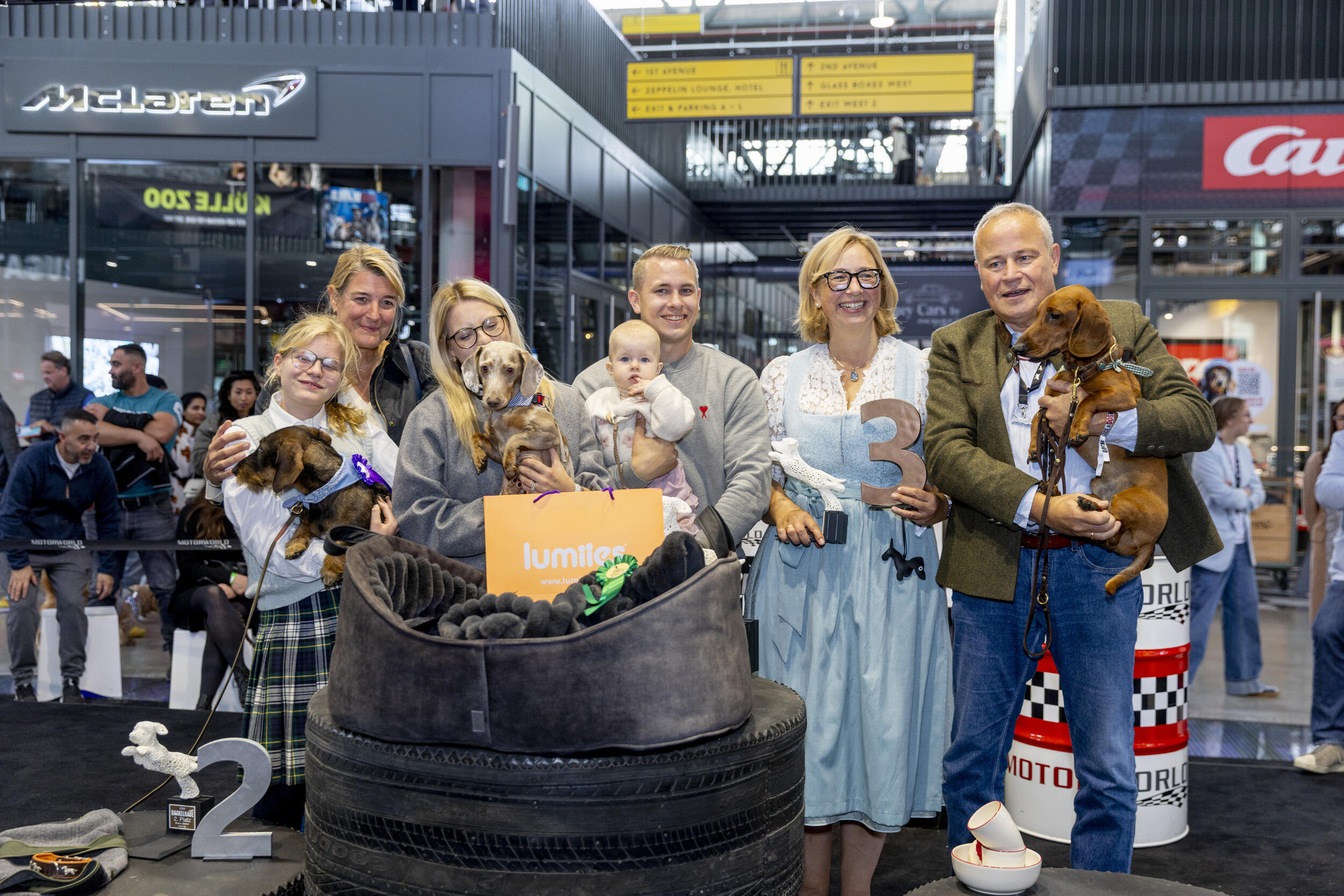 Gruppenfoto von sieben Menschen, darunter ein Baby und mehrere Frauen in traditioneller Kleidung, mit drei Dackelhunden auf einem Podium aus Autoreifen bei einer Veranstaltung in einer Halle mit McLaren-Logo und Wegweiserschildern im Hintergrund.