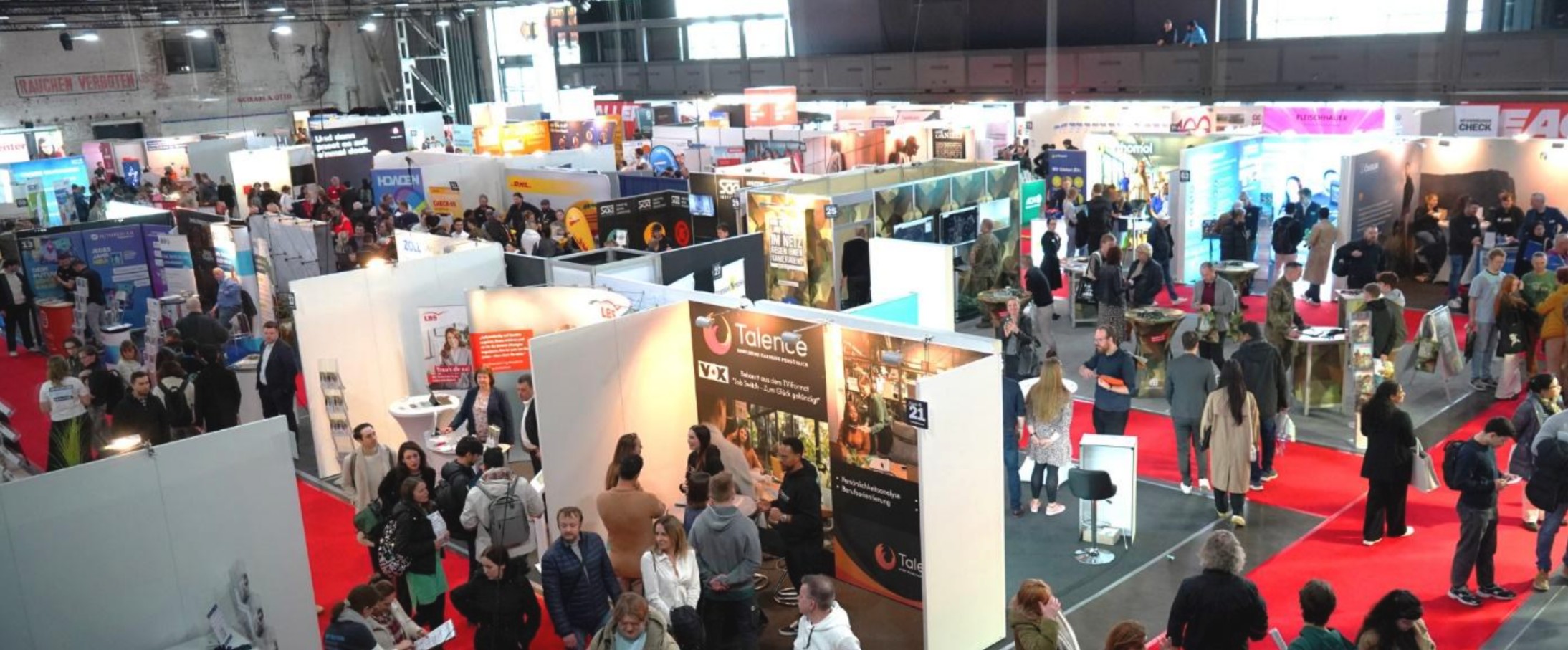 Blick von oben auf eine belebte Messehalle mit mehreren Ständen, vielen Besuchern und roten Teppichwegen, zahlreiche Menschen unterhalten sich und besichtigen die Ausstellungsflächen.