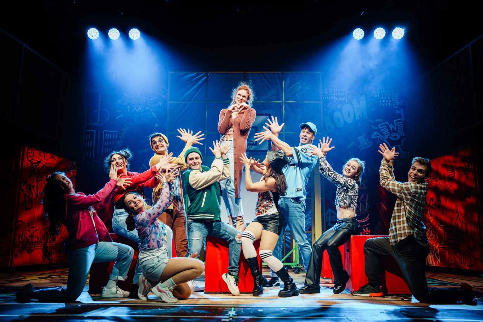 Eine Theatergruppe auf der Bühne, bestehend aus elf jungen Darstellern in bunter moderner Kleidung, die eine fröhliche, ausdrucksstarke Pose machen, wobei eine Person in der Mitte auf einem roten Kasten steht und die anderen sie umgeben und mit den Händen auf sie zeigen, beleuchtet von blauem Bühnenlicht mit Graffiti-ähnlichem Hintergrund.