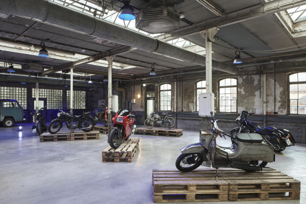 Eine industrielle Ausstellungshalle mit mehreren Oldtimer-Motorrädern auf Holzpaletten, wobei im Vordergrund ein silberner Motorroller steht, im Hintergrund weit geöffnete Fenster und eine blaue alte Kastenwagen-Lieferfahrzeug