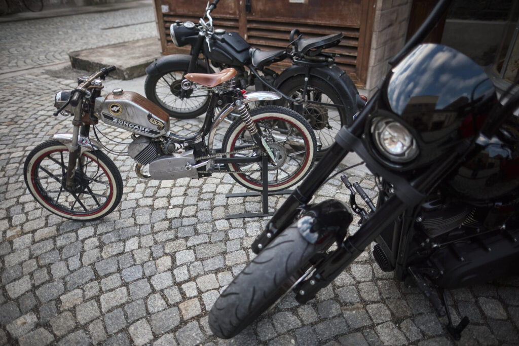 Drei Motorräder unterschiedlicher Stile auf Kopfsteinpflaster, vorne ein modernes schwarzes Motorrad mit rundem Scheinwerfer, in der Mitte ein klassisches Zündapp-Moped mit braunem Sitz und Tank, hinten ein schwarzes altes Motorrad.